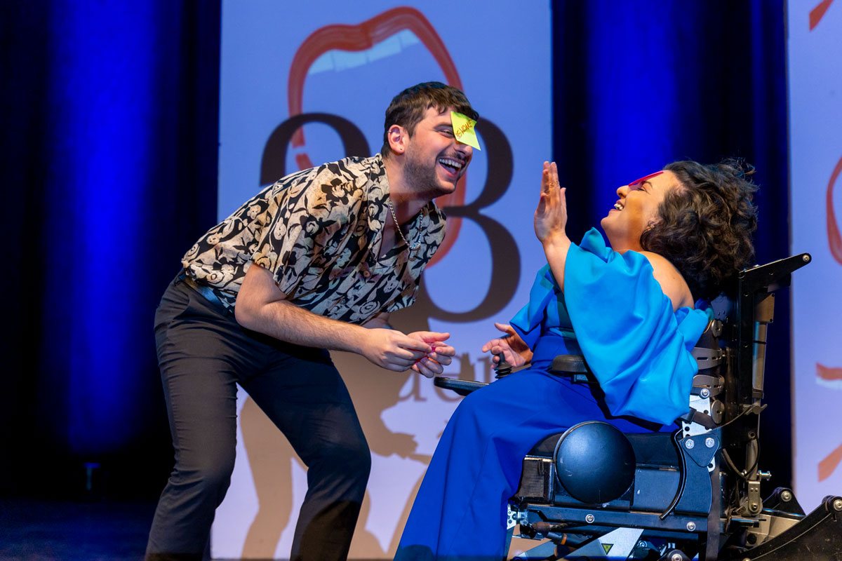 Eine Schauspielerin und ein Schauspieler lachen auf einer Bühne und geben sich ein High-Five. Die Schauspielerin im blauen Oberteil sitzt im Rollstuhl, der Schauspieler steht in einem schwarz-weiß gemusterten Hemd daneben und hat einen gelben Klebezettel 
