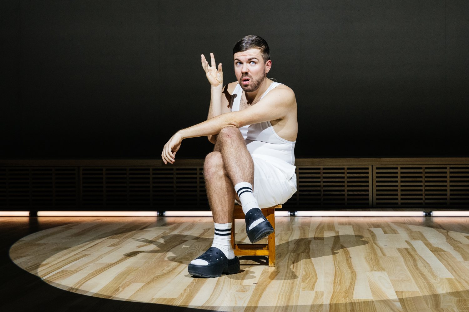 Eine Person sitzt auf einem kleinen Holzhocker auf einer leeren Bühne. Sie hat kurze Haare und einen Bart, trägt ein weißes Tanktop, weiße Shorts, weiße Socken mit schwarzen Ringeln und klobige schwarze Sandalen. Mit aufgerissenen Augen und einer erhobene