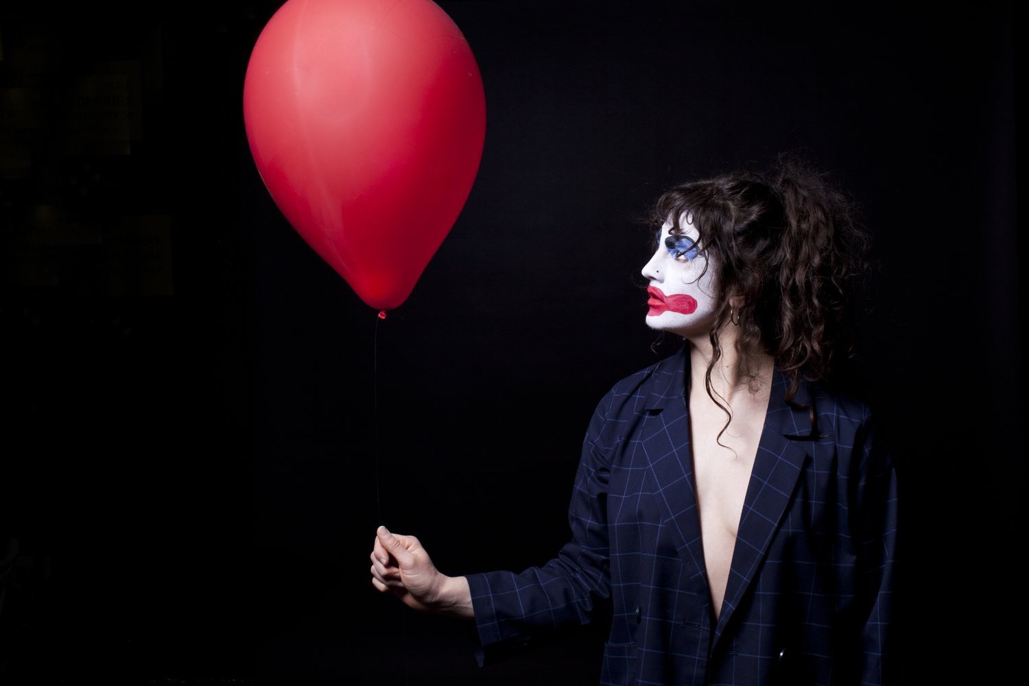 Eine Performer*in im Profil vor schwarzem Hintergrund mit weiß-blau-rotem Clown-Make-up. Sie trägt ein dunkles, kariertes Sakko über dem nackten Oberkörper und hält die Schnur eines roten Luftballons in der Hand.