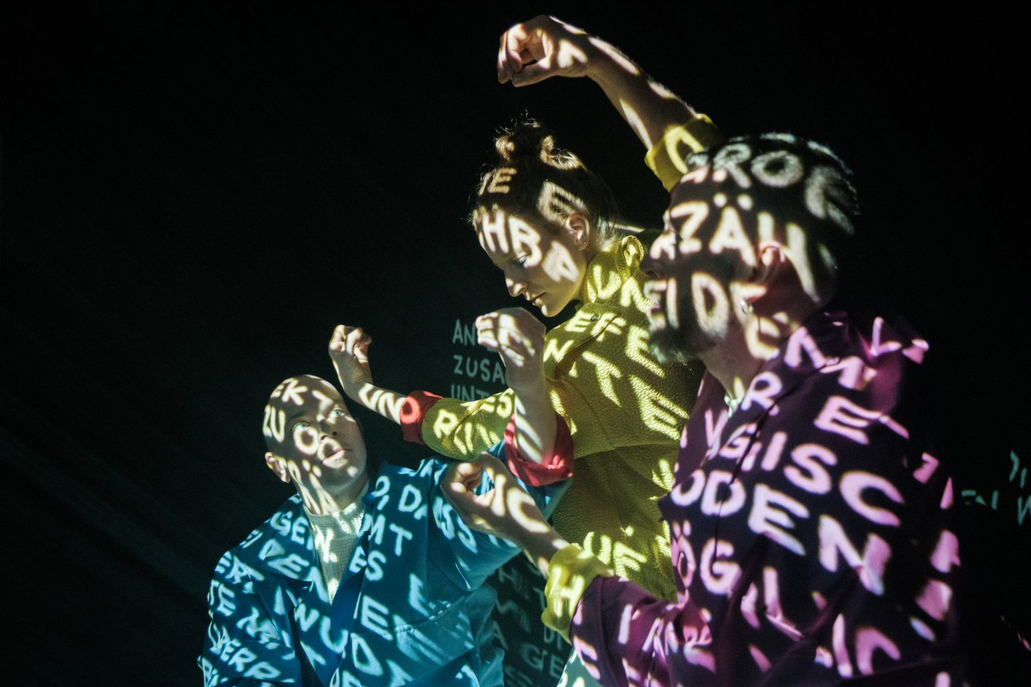 Nahaufnahme von drei Performer*innen vor schwarzem Hintergrund. Sie gestikulieren mit den Händen, während ihre Gesichter und Oberkörper von einer Lichtprojektion aus vielen weißen Buchstaben und Textfragmenten angestrahlt werden.