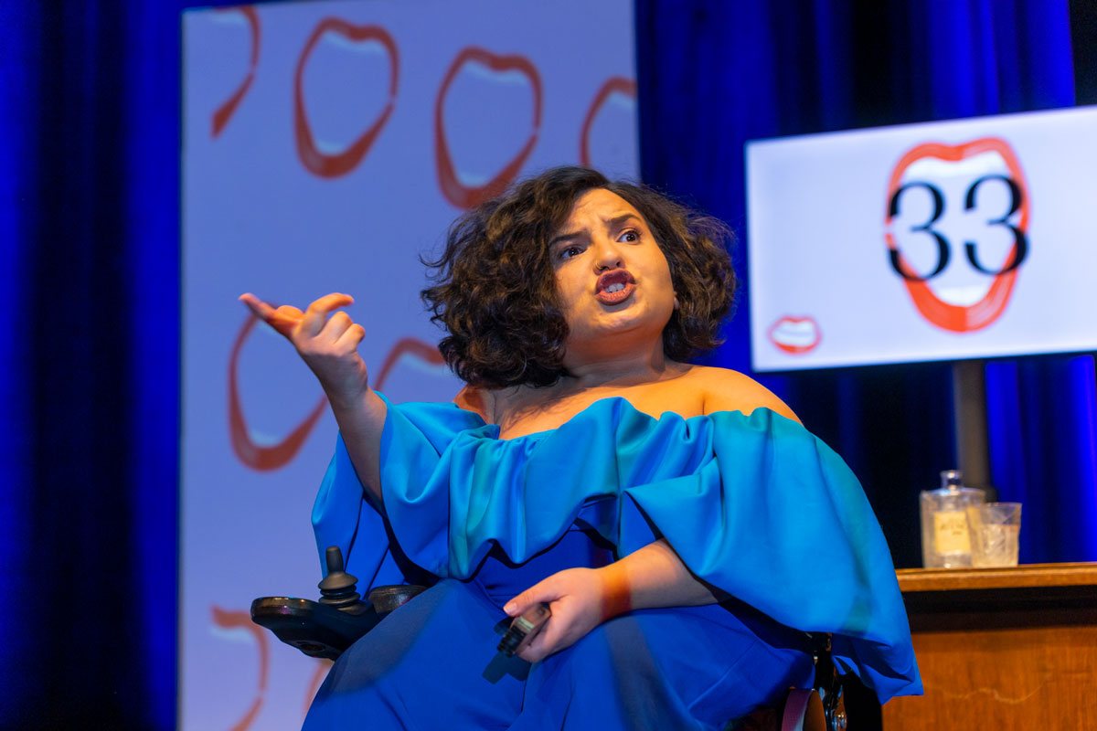 Eine Schauspielerin mit dunklen Locken und leuchtend blauem, schulterfreiem Oberteil sitzt im Rollstuhl auf einer Bühne und spricht gestikulierend. Im Hintergrund zeigt ein Bildschirm die Zahl 33 innerhalb eines rot gezeichneten Mundes.