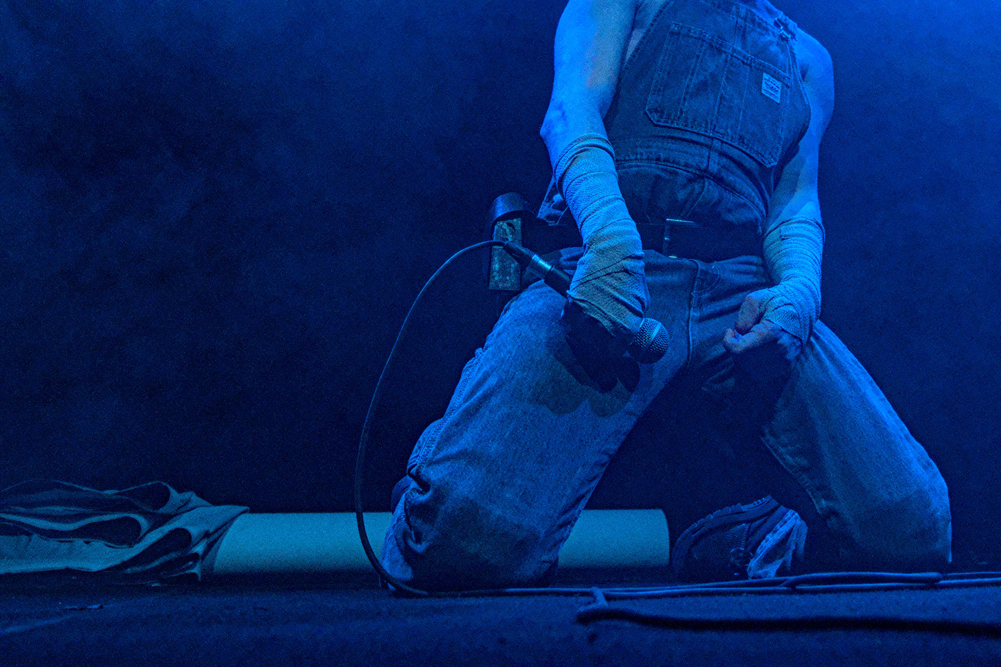 Ein kniender Performer in Latzhose unter starkem blauen Scheinwerferlicht. Die Unterarme und Hände sind fest in Bandagen gewickelt und umgreifen ein Mikrofon.
