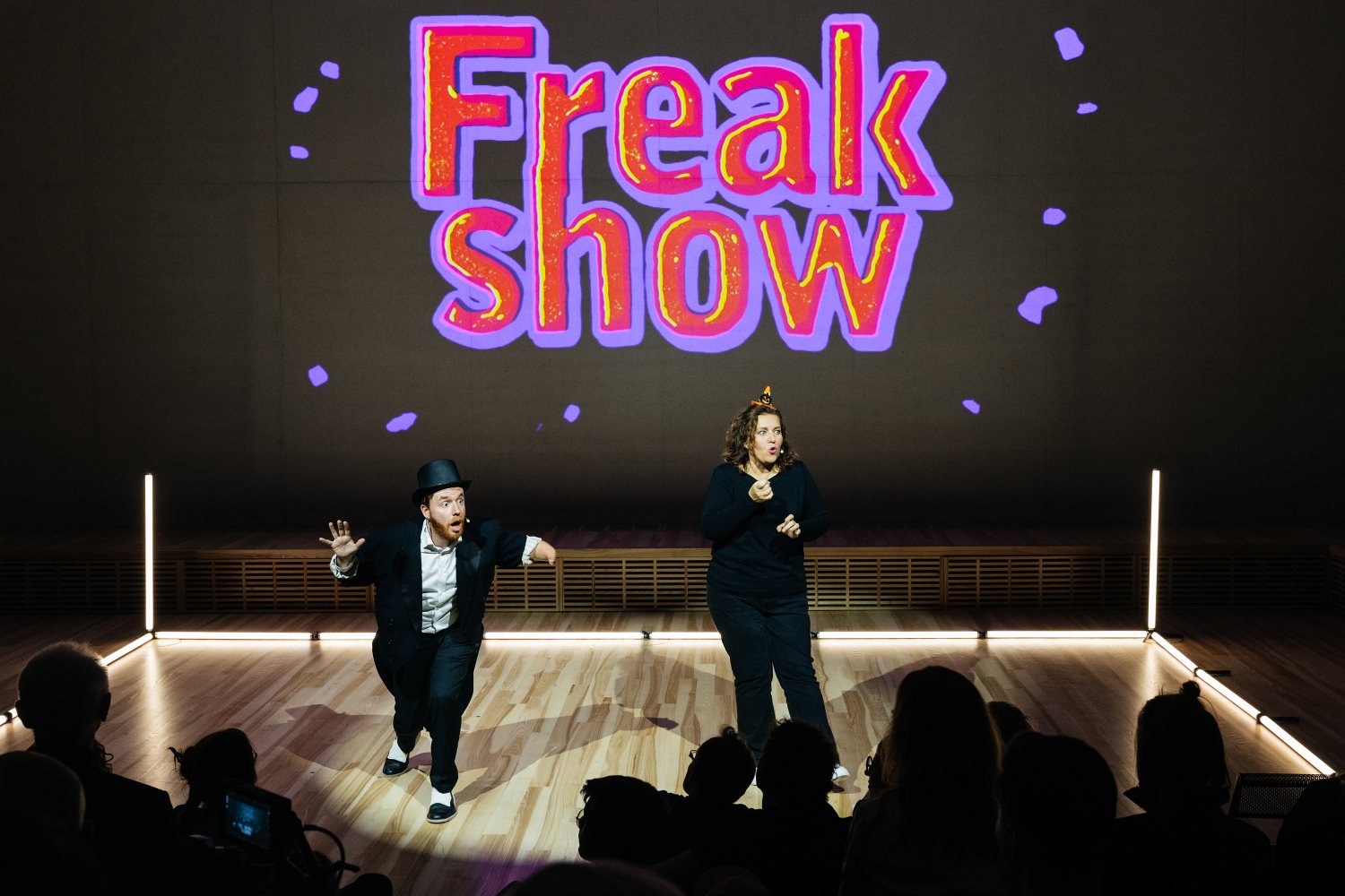 Eine Theaterbühne mit einer großen, bunten Projektion des Wortes „Freakshow“. Vorne agieren zwei Darsteller*innen: Eine Person im schwarzen Anzug mit Zylinder beugt sich dynamisch vor, daneben steht eine Person in dunkler Kleidung und gestikuliert mit den