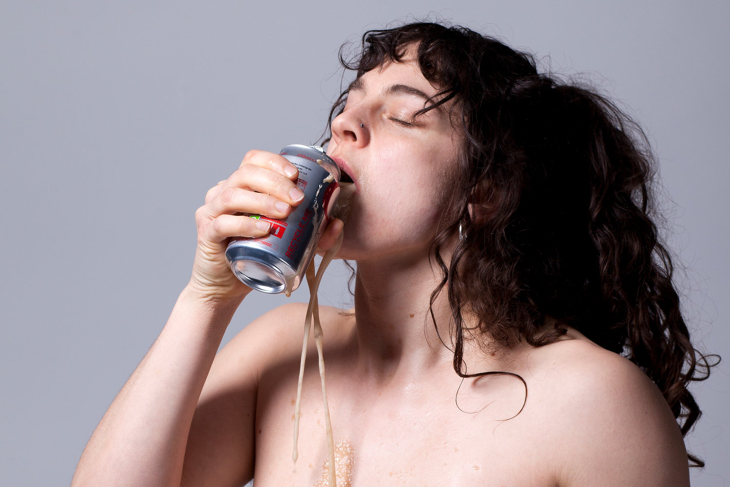 Porträt einer Performer*in mit geschlossenen Augen und freiem Oberkörper, die hastig aus einer silbernen Dose trinkt, wobei die helle Flüssigkeit aus dem Mund über Kinn und Brust herabläuft.