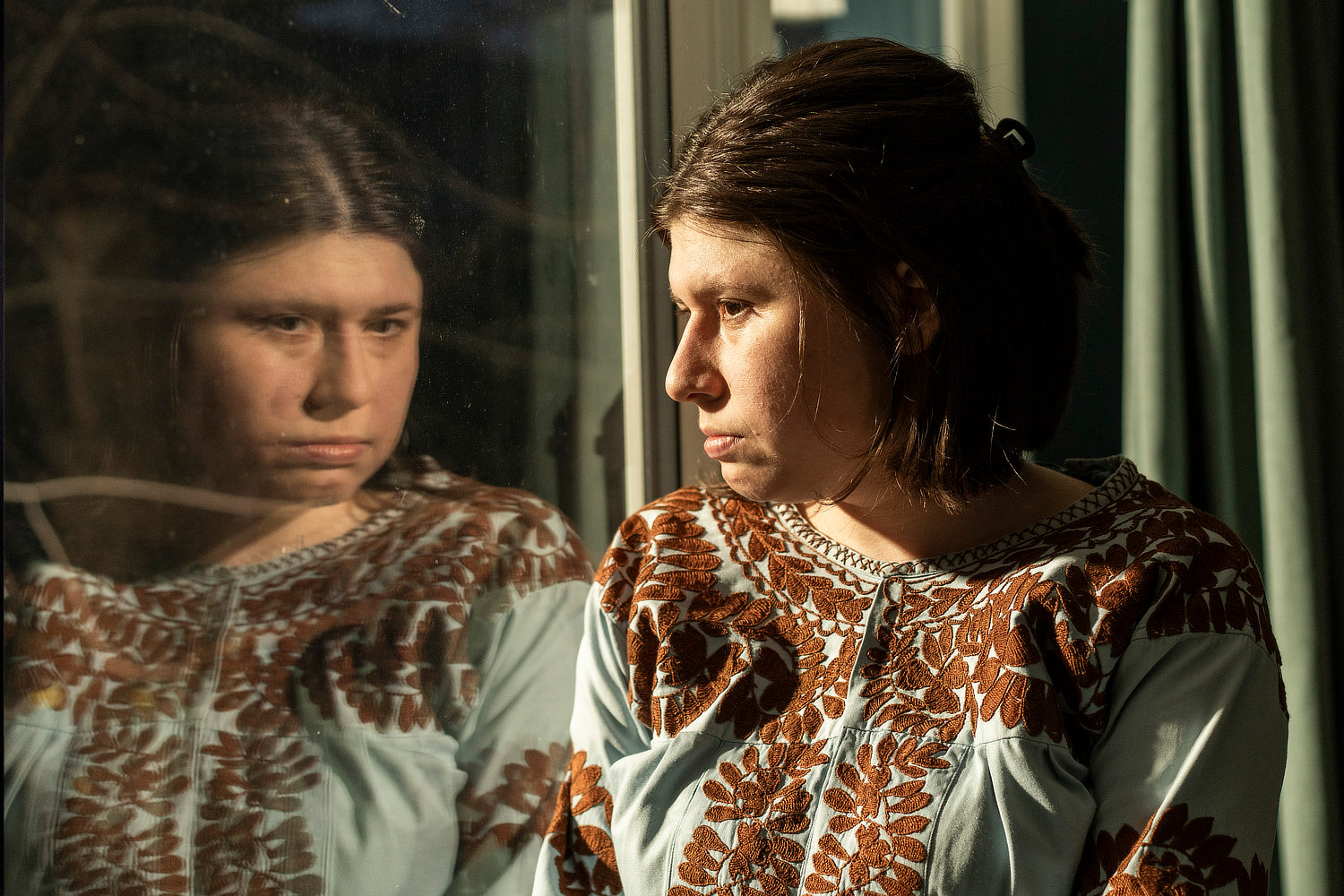 Eine junge Frau in einer hellblauen Bluse mit brauner Blumenstickerei steht im Sonnenlicht und blickt nachdenklich aus einem Fenster. Ihr Gesicht spiegelt sich in der Glasscheibe.