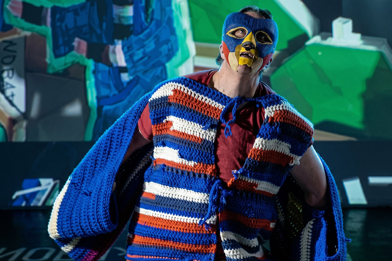 Nahaufnahme einer Person mit blau-gelber Maske und einem grob gehäkelten, bunt gestreiften Oberteil. Die Person blickt mit leicht geöffnetem Mund nach oben. Im Hintergrund sind unscharfe, abstrakte geometrische Formen in leuchtenden Farben zu sehen.