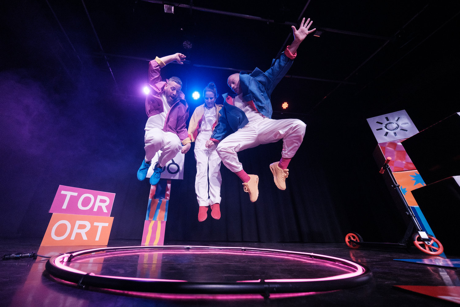 Drei Performer:innen in weißen Outfits und bunten Jacken springen energiegeladen auf einer dunklen Bühne in die Luft. Auf dem Boden liegen ein pink leuchtender Ring, ein Tretroller und bunte Klötze mit den Wörtern "TOR" und "ORT".