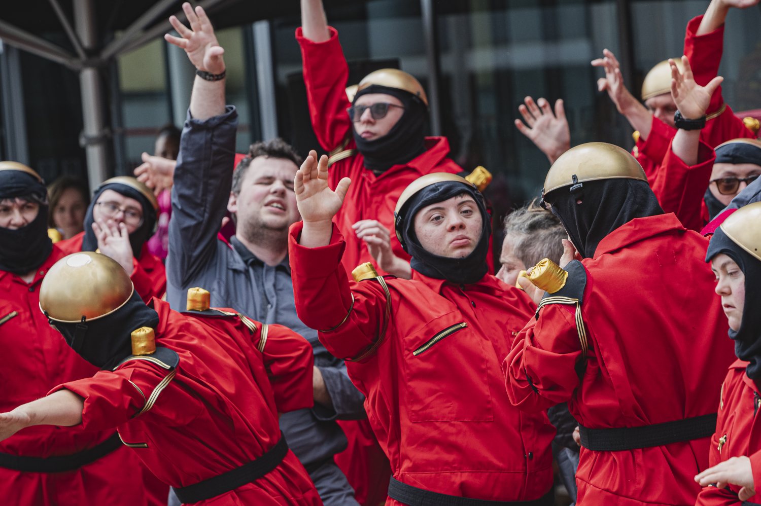 Nahaufnahme einer Gruppe von Tänzer*innen im Freien. Die meisten tragen rote Overalls sowie goldene Helme und Handschuhe und haben die Arme in die Luft gestreckt. Zwischen ihnen steht eine Person in einem graublauen Overall.