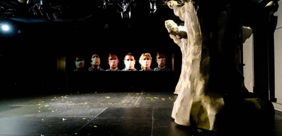 Auf die Bühnenrückwand sind sechs Personen projiziert. Sie tragen alle die selben Hemden und Mundschutz. Am vorderen Bühnenrand steht ein knorriger Baum.