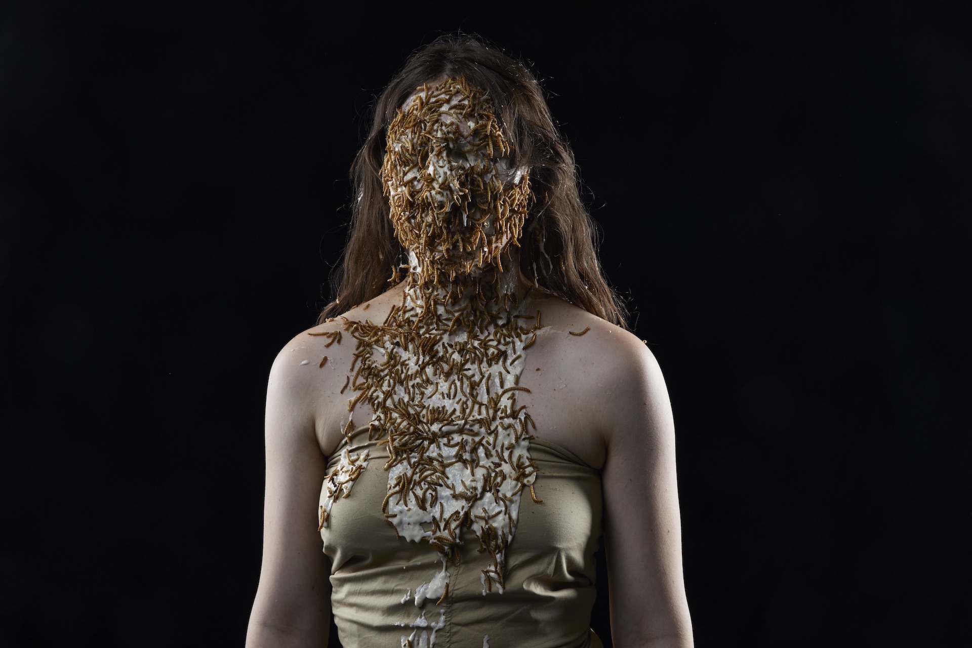 Eine junge Frau mit langem, braun gewelltem Haar und goldenem Bustier vor schwarzem Grund. Ihr gesamtes Gesicht und Teile des Dekolleté ist mit Maden bedeckt.