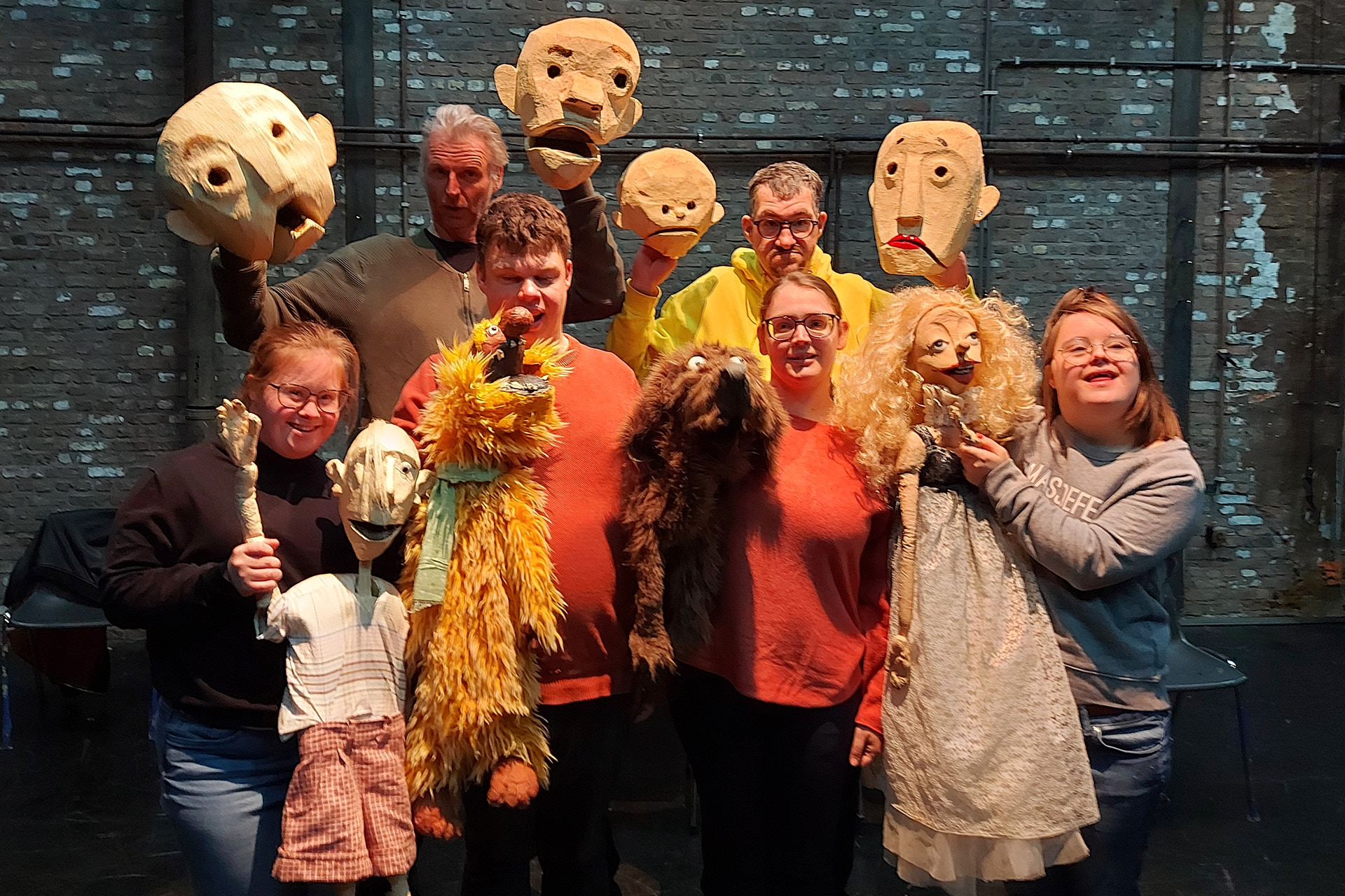 Sechs Personen in Alltagskleidung als Gruppe vor einer blau gekachelten Wand. Sie halten verschiedene Puppen aus Fell oder große runde Gesichtsmasken in die Höhe.